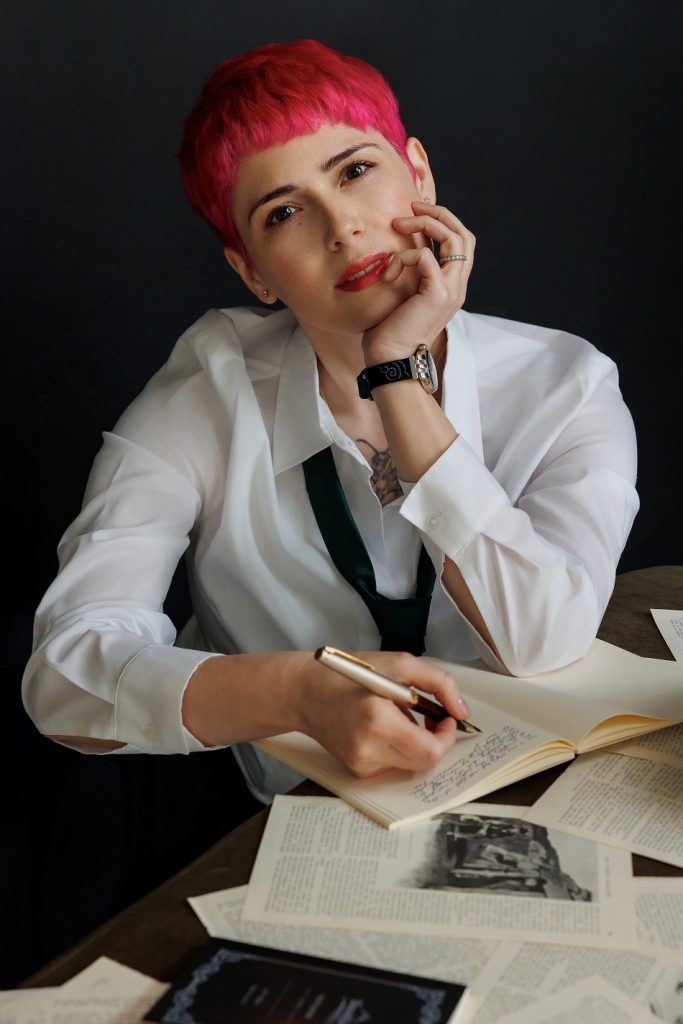 Picture of pink-haired white woman in white shirt with loose tie, hints of a tattoo peaking out of her shirt, writing with a fountain pen in a journal.